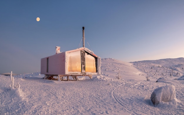 Maison rouge solitaire sur une colline enneigée et givrée sous le ciel du lever du soleil avec une pleine lune Arrière-plan minimaliste panoramique avec une cabane rouge solitaire en hiver Confort des vacances d'hiver par une nuit glaciale