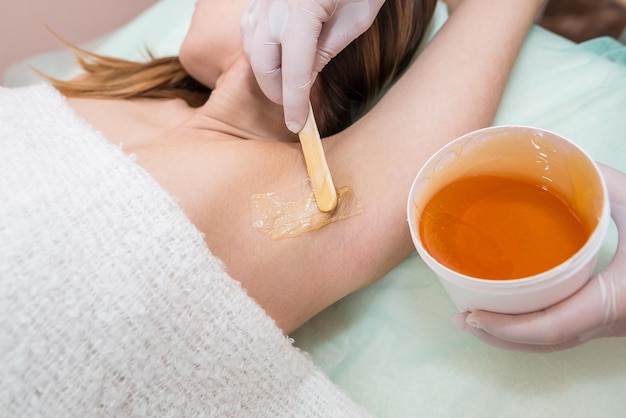 Le maître applique de la pâte à sucre sur l'aisselle d'une jeune femme pour enlever les cheveux Épilation des aisselles Shugaring avec du sucre liquide Concept de beauté