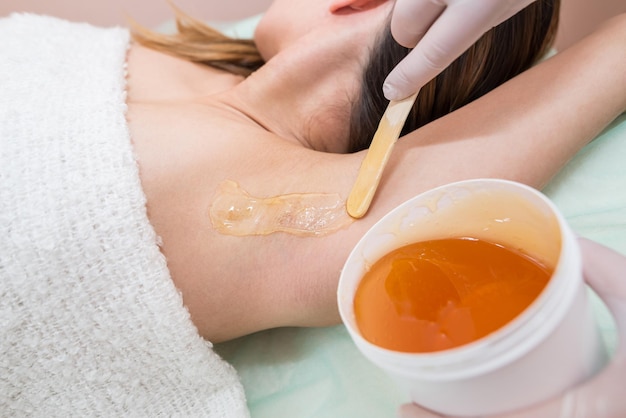 Le maître applique de la pâte à sucre sur l'aisselle d'une jeune femme pour enlever les cheveux Épilation des aisselles Shugaring avec du sucre liquide Concept de beauté
