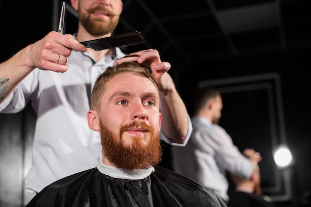 Le maître coupe les cheveux et la barbe dans le salon de coiffure. Le coiffeur fait la coiffure à l'aide de ciseaux et d'un peigne en métal