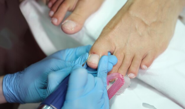 Maître en gants faisant une pédicure sur les ongles de pied dans un salon de beauté en utilisant un gros plan de forage