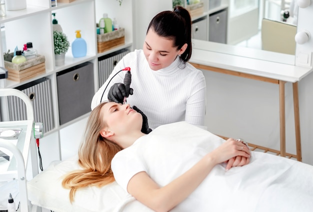 Maître en masque de médecine tenant une machine à encre et faisant un maquillage permanent des sourcils. Microblading en temps de covid