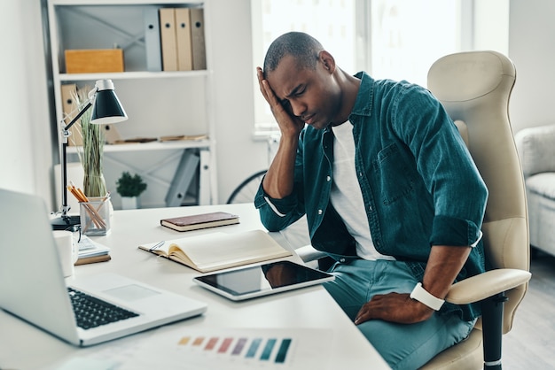 Mal de crâne. Jeune homme africain frustré en chemise faisant une grimace alors qu'il était assis au bureau