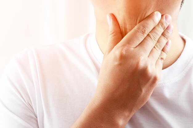 Mal de gorge. Gros plan d'un jeune homme malade tenant sa gorge enflammée à l'aide des mains pour toucher le cou malade en chemise bleue sur fond gris. Concept médical et de soins de santé. Concentrez-vous sur le rouge pour montrer la douleur.