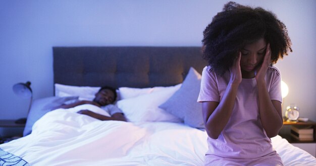 Ce mal de tête m'a tenu éveillé toute la nuit Photo d'une jeune femme inquiète assise sur un lit avec son mari dormant en arrière-plan