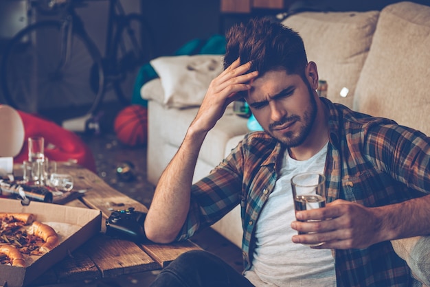 Ce mal de tête me tue ! Beau jeune homme tenant un verre d'eau et ayant l'air épuisé