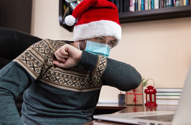 Maladie de Noël. Jeune homme en chapeau de père Noël et masque facial tousse dans le coude et parle à l'aide d'un ordinateur portable pour les amis et les enfants des appels vidéo. Noël pendant le coronavirus.