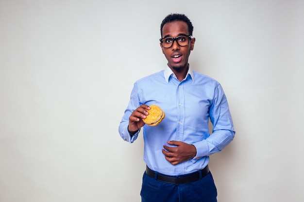 Maladie triste homme afro-américain a mis la main sur l'abdomen de la douleur de hamburger.beau et jeune homme afro dans une chemise élégante et des lunettes tenant un hamburger sur fond blanc. malbouffe diète indigestion.