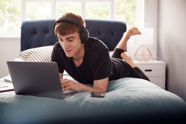 Male College Student Wearing Headphones se trouve sur le lit dans une maison partagée travaillant sur un ordinateur portable