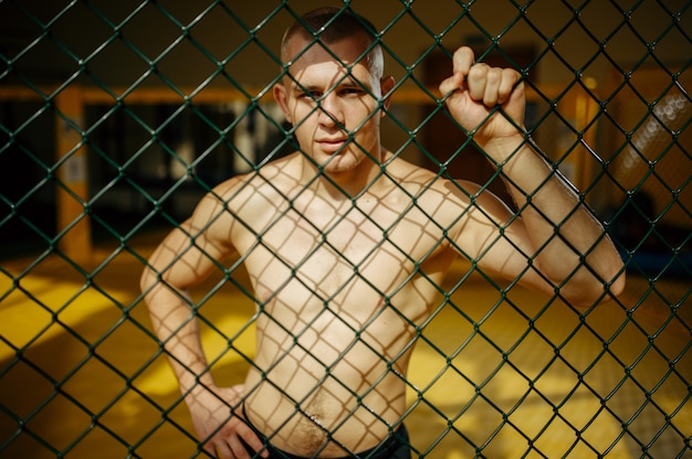 Mâle combattant MMA debout à la grille dans une cage dans une salle de sport.