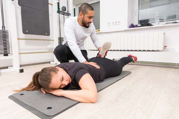 Mâle formateur étirement veau de jeune femme allongée sur le tapis dans la salle de gym.