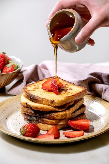 Mâle main verser le sirop d'érable à partir de la cruche en céramique sur le stock de pain grillé français avec des fraises fraîches sur plaque et serviette en tissu rose sur tableau blanc