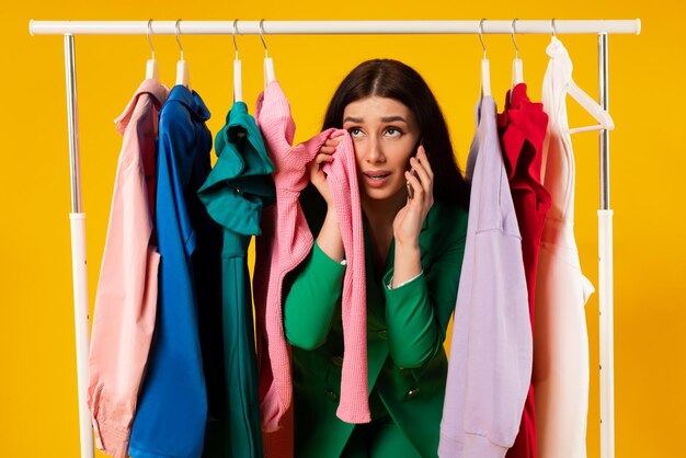 Photo malheureuse femme accro du shopping pleurant et parlant au téléphone portable debout près du rail de vêtements sur fond jaune