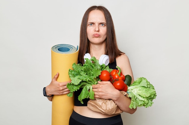 Malheureuse femme de race blanche tenant des légumes biologiques debout isolé sur fond blanc regardant la caméra avec des lèvres moue expression offensée a séance d'entraînement sur tapis