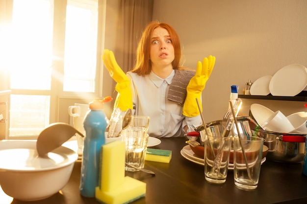 Malheureuse gingembre roux belle femme portant un gant jaune et lave des montagnes de tas de vaisselle sale : tasses, casserole, casserole dans la cuisine.