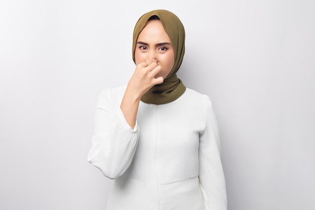 Malheureuse jeune belle femme musulmane asiatique des années 20 portant le hijab se sentant dégoûtée tenant son nez pour éviter de sentir l'appareil photo fétide isolé sur fond blanc Les gens concept de style de vie religieux