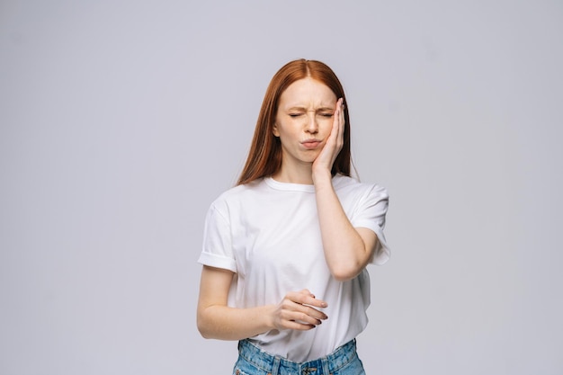 Malheureuse jeune femme bouleversée touche la joue souffrant d'une sensation soudaine de douleur dentaire sur fond blanc isolé. Jolie mannequin aux cheveux roux montrant émotionnellement des expressions faciales, espace de copie