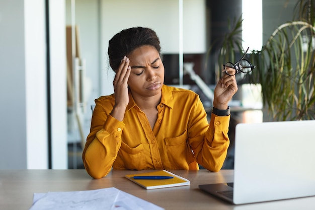 Malheureuse pigiste noire se sentant fatiguée de travailler sur un ordinateur portable souffrant de maux de tête à l'intérieur du bureau