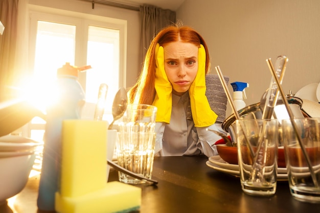 Malheureux gingembre roux belle femme portant des gants jaunes et laver des tas de montagnes de vaisselle sale cupspansaucepan dans la cuisine