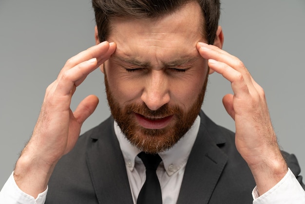 Malheureux homme barbu frustré portant un costume de style officiel massant les tempes ressentant des maux de tête souffrant de migraine ou d'hypertension artérielle tourné en studio intérieur isolé sur fond gris