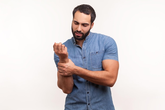 Malheureux homme barbu malade en chemise bleue touchant la main douloureuse, souffrant d'un traumatisme, d'une entorse au poignet, ressentant la douleur du syndrome du canal carpien. Tourné en studio intérieur isolé sur fond blanc