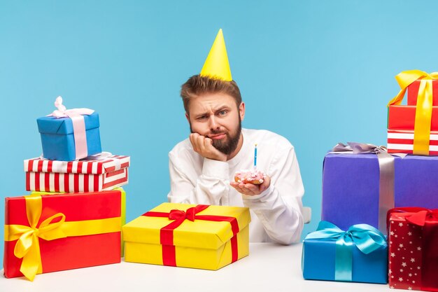 Malheureux homme ennuyé en chapeau conique tenant un gâteau avec une bougie assis sur le lieu de travail avec de nombreuses boîtes-cadeaux décorées solitaire célébrant son anniversaire au travail Studio intérieur tourné isolé sur fond bleu