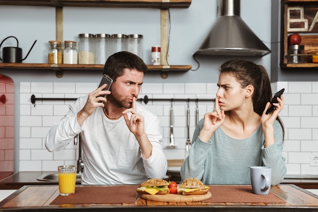 Malheureux jeune couple assis à la cuisine pendant le déjeuner à la maison, ayant un problème, tenant un téléphone mobile