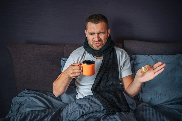 Malheureux jeune homme est assis sur le lit dans la chambre et regarde la tasse orange. Il le tient dans une main et un morceau de citron dans une autre. Guy n'est pas satisfait. Il se rétrécit. Le liquide dans la tasse sent dégoûtant.