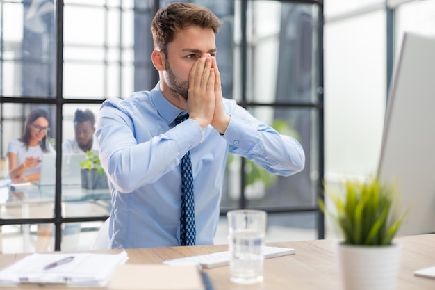 Malheureux jeune homme frustré tenant la tête par les mains assis avec un ordinateur derrière le bureau au bureau avec des collègues en arrière-plan