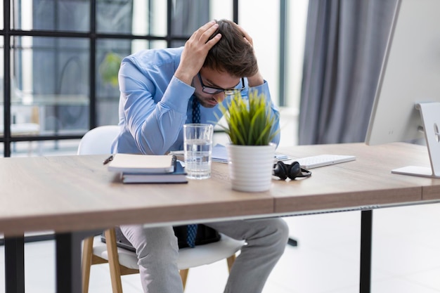 Malheureux jeune homme frustré tenant la tête par les mains assis avec un ordinateur derrière le bureau au bureau