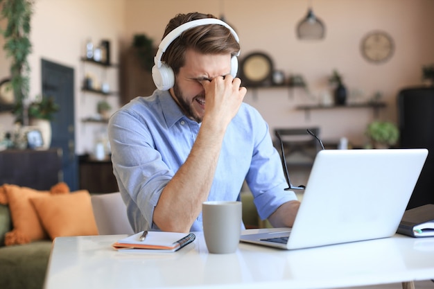 Malheureux jeune homme frustré tenant la tête par les mains assis avec un ordinateur portable derrière le bureau à la maison.