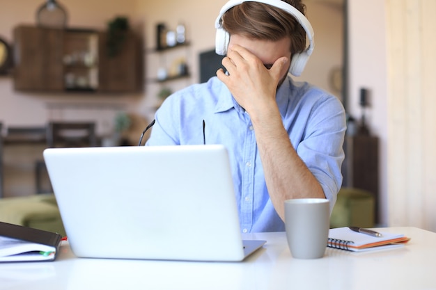 Malheureux jeune homme frustré tenant la tête par les mains assis avec un ordinateur portable derrière le bureau à la maison.