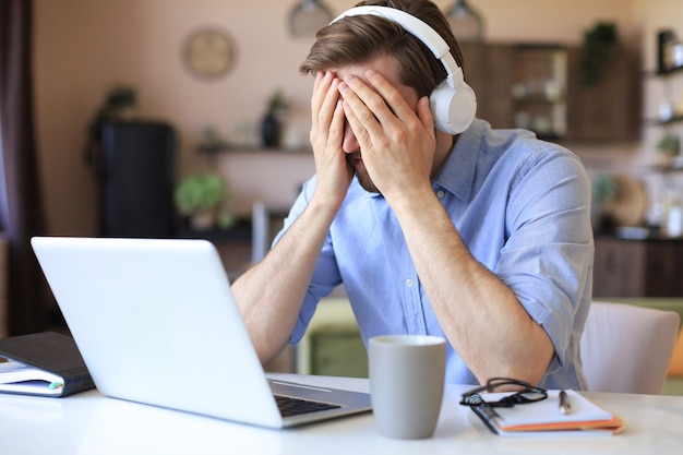 Malheureux jeune homme frustré tenant la tête par les mains assis avec un ordinateur portable derrière le bureau à la maison.