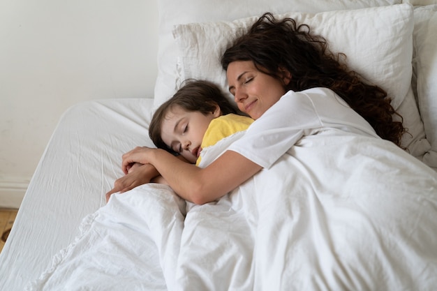 Maman aimante paisible embrasse son fils d'âge préscolaire dormant avec un enfant dans un lit confortable sous une couverture chaude