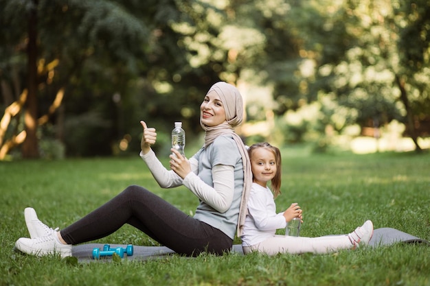 Maman arabe et sa fille assises sur un tapis de transport gris dans le parc d'été tenant des bouteilles d'eau