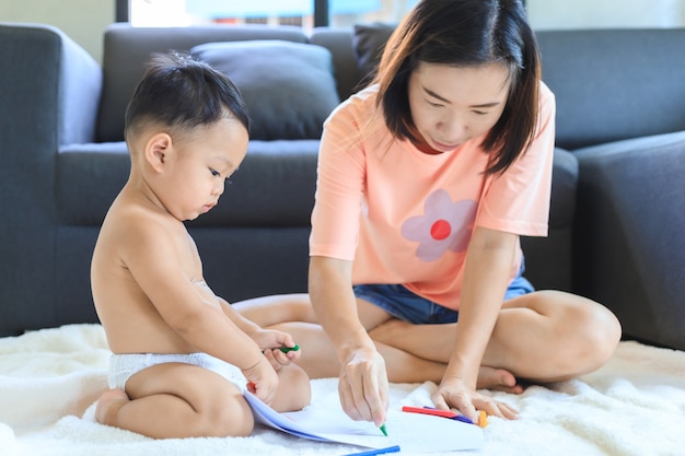 Photo maman asiatique apprend à son mignon petit garçon à utiliser des crayons de couleur pour dessiner sur papier à la maison. concept de famille et de convivialité.