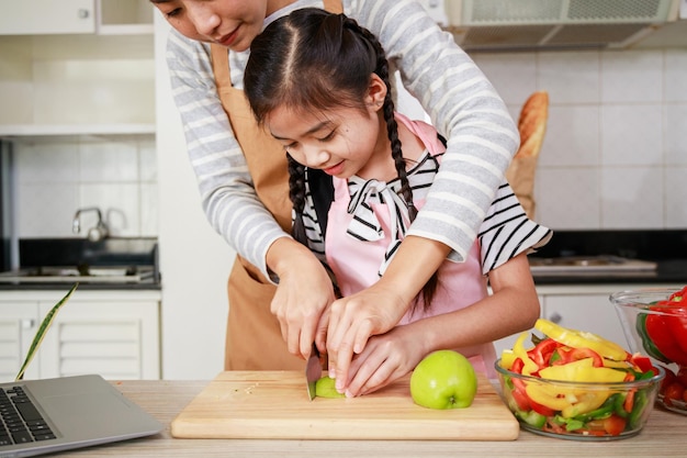 Maman asiatique et enfant fille tranchent la pomme verte ensemble pour préparer une salade de légumes dans la cuisine