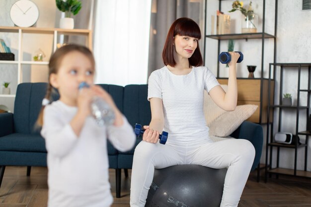 Maman assise sur un ballon de fitness et faisant des exercices sportifs avec des haltères avec sa petite fille