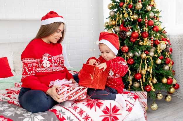 Maman et bébé dans un pull rouge et des chapeaux offrent des cadeaux sous le sapin de Noël en se félicitant à la maison sur le lit et se réjouissent de la nouvelle année et de Noël