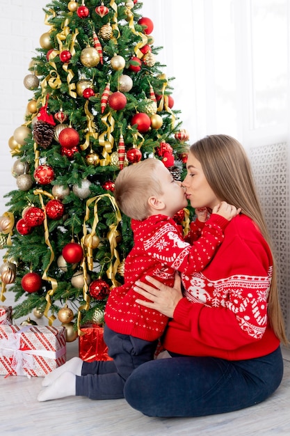 Maman et bébé dans un pull rouge sous le sapin de Noël à la maison profitent de la nouvelle année et de Noël, s'embrassent et se félicitent