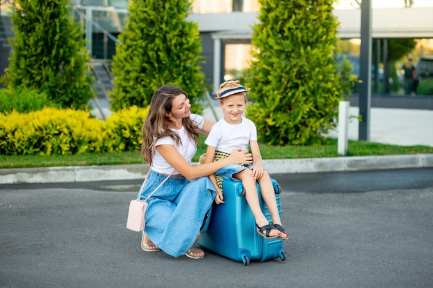 Une maman et un bébé heureux sont assis sur une valise bleue devant l'aéroport et partent en vacances ou se rendent à l'aéroport