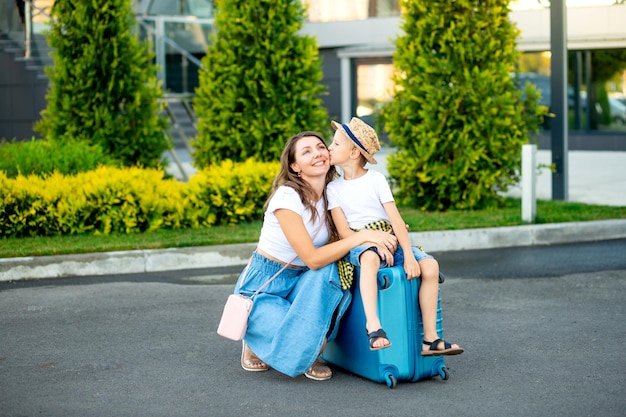 Une maman et un bébé heureux sont assis sur une valise bleue devant l'aéroport et partent en vacances ou se rendent à l'aéroport