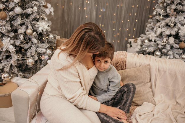 Une maman blonde est assise sur le canapé et serre un petit garçon dans ses bras sur un fond avec un sapin de Noël