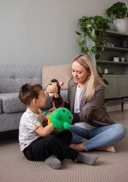 Une maman blonde est assise sur le tapis dans la chambre et joue au théâtre de marionnettes avec son fils