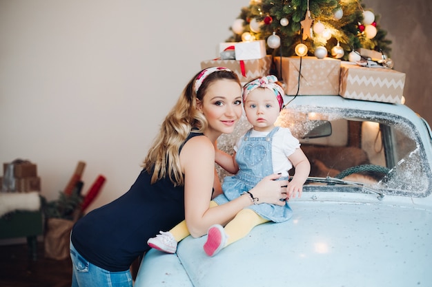 Maman blonde à la mode avec petite fille mignonne posant ensemble, assis sur une voiture rétro bleue.