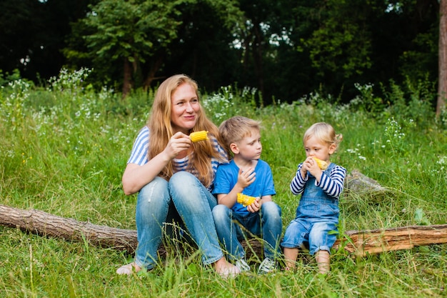 Maman avec deux enfants assis dans les bois sur un tronc d'arbre tombé et mangeant du maïs