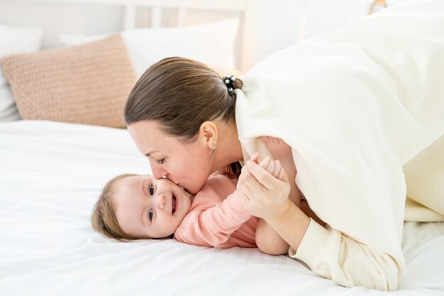 Maman embrasse doucement et serre dans ses bras sa petite fille à la maison sur le lit et s'amuse ensemble la maternité familiale et l'amour pour le bébé