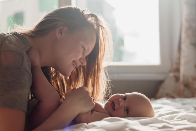 Maman embrasse et embrasse un nouveau-né souriant allongé sur le lit jeune femme avec un enfant à la maison