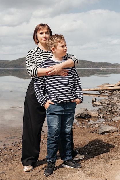 Photo maman embrasse son fils au bord de la rivière. une belle mère moderne avec un fils heureux dans des gilets rayés.