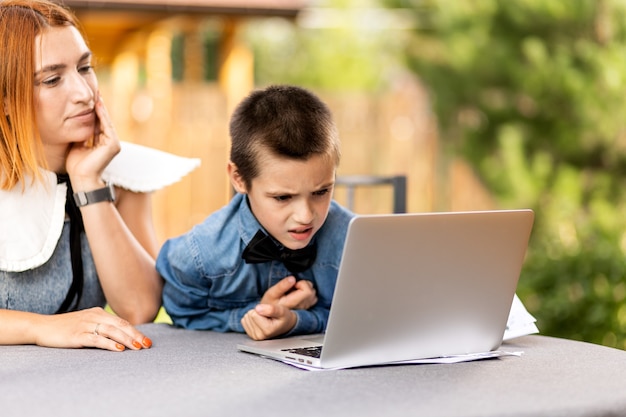 Maman enseigne à l'enfant à la maison dans le jardin, l'enseignement à domicile. Maman aide son fils à faire ses devoirs, le stress. Cours scolaires en ligne via ordinateur portable